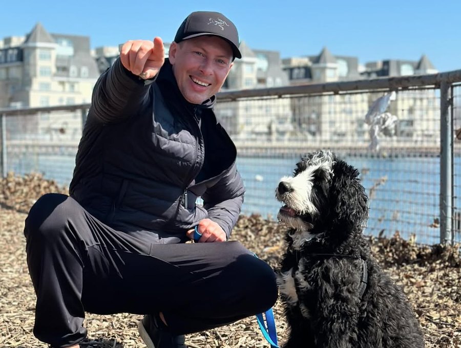 A walker crouching by the water, grinning beside a bernedoodle on a one-to-one walk